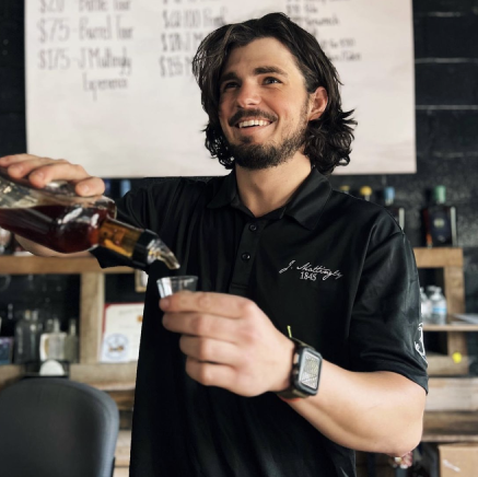 A bartender pouring bourbon in a shot glass for tasting and smiling.